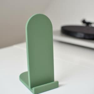 Op de afbeelding: Een saliegroene telefoonstandaard met een boogvormig ontwerp en een gladde, matte afwerking. De standaard heeft een verticaal achterpaneel en een kleine horizontale richel aan de basis om een mobiel apparaat rechtop op een vlak te houden.