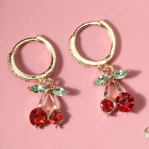 Puede incluir: Pendientes de aro dorados con un colgante de cereza. La cereza está hecha de piedras de cristal rojas y verdes.