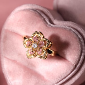 May include: A gold ring with a pink flower design featuring clear and pink gemstones. The ring is displayed in a pink velvet heart-shaped box.