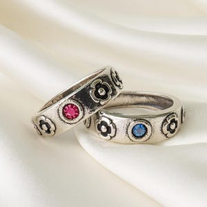 May include: Two silver rings with flower designs and colored gemstones. One ring features a pink gemstone, the other a blue gemstone. The rings are set against a white, silky background.
