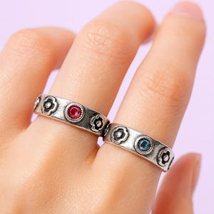 May include: Two silver rings with a floral design. One ring features a red gemstone, the other a blue gemstone. The rings are worn on fingers against a light purple background. The rings have a vintage, antique style.