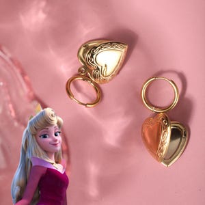 May include: A pair of gold hoop earrings with a heart-shaped locket dangling from each hoop. The lockets have a detailed design and are open to reveal a mirror inside.
