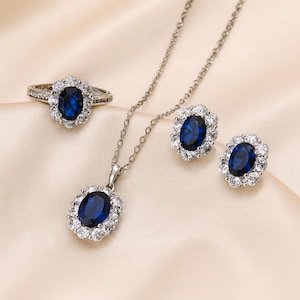 Op de afbeelding: Een sieradenset met een ring, hanger en oorbellen. Elk stuk heeft een grote, ovale, donkerblauwe edelsteen omringd door kleinere, heldere stenen. De ring en hanger hangen aan een fijne zilveren ketting.