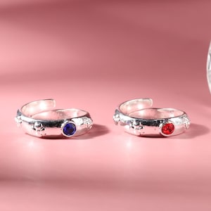 May include: Two silver toe rings with adjustable bands. One ring has a blue gemstone and the other has a red gemstone.