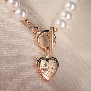 May include: A pearl necklace with a gold-colored clasp and a heart-shaped pendant. The pendant has the words "I Love You" engraved on it, along with floral details. The necklace is made of round, white pearls.