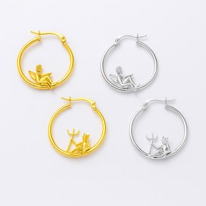 May include: Gold and silver-toned hoop earrings. Each earring features a small figure inside the hoop. Two gold earrings have a figure with wings, and two have a figure with a trident. The silver earrings have the same figures.