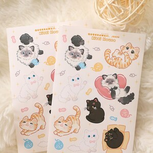 May include: Two sheets of stickers featuring various cartoon cat illustrations. The stickers include cats in different poses, colors, and patterns, with some holding yarn balls or wearing bows. The text "QUOKKAWAII. Kitti House" is printed on the stickers.
