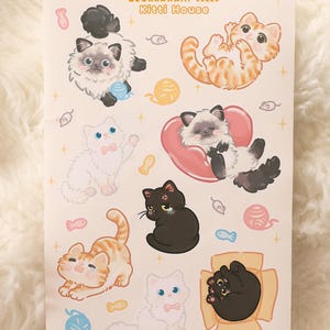 May include: A sheet of stickers featuring cartoon cats in various poses. The stickers include cats in orange, black, and white, with some wearing bows or resting on hearts. The text "Studio Quokkawaii. J0226 Kitti House" is at the top.