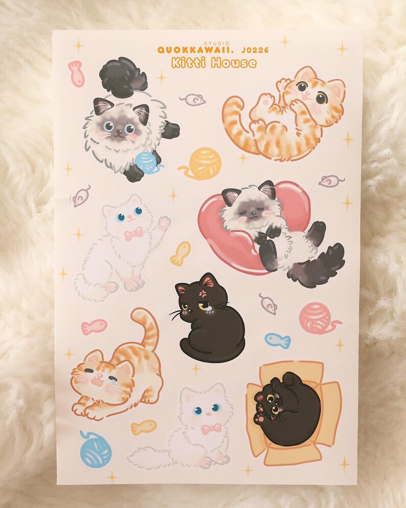 May include: A sheet of stickers featuring cartoon cats in various poses. The stickers include cats in orange, black, and white, with some wearing bows or resting on hearts. The text "Studio Quokkawaii. J0226 Kitti House" is at the top.