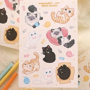 May include: A sheet of stickers featuring various cartoon cats in different poses. The cats are in shades of black, white, and orange, with some wearing pink bows or resting on pink heart-shaped pillows. The text "QUOKKAWAII. J0226 Kitti House" is printed on the sheet.