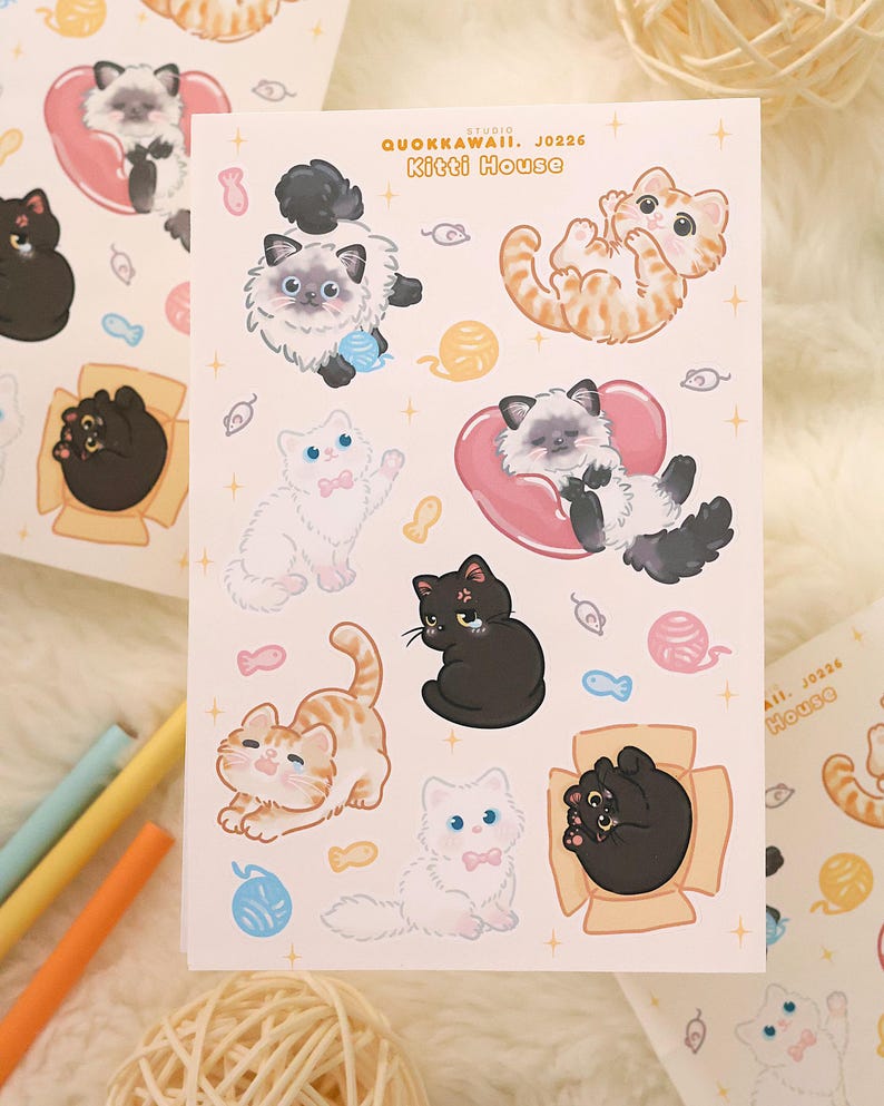 May include: A sheet of stickers featuring various cartoon cats in different poses. The cats are in shades of black, white, and orange, with some wearing pink bows or resting on pink heart-shaped pillows. The text "QUOKKAWAII. J0226 Kitti House" is printed on the sheet.