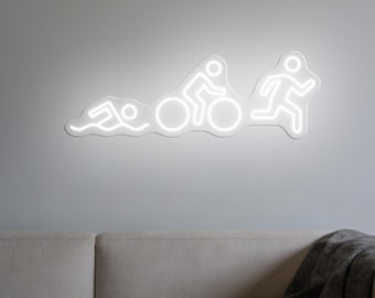 May include: A white neon sign depicting the stages of a triathlon: swimming, cycling, and running. The sign is mounted on a gray wall above a light-colored sofa with a gray blanket.