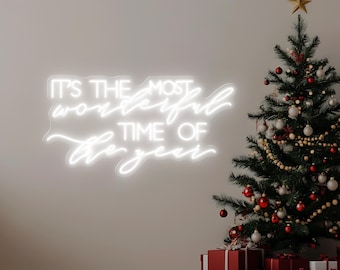 May include: A white neon sign on a wall reads "IT'S THE MOST WONDERFUL TIME OF THE YEAR." A decorated Christmas tree with red and white ornaments, a gold star, and wrapped gifts are in the foreground. The gifts are wrapped in red and white paper.