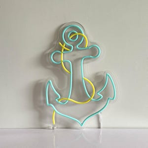 Puede incluir: Una l&aacute;mpara de ne&oacute;n con forma de ancla. El ancla es de color azul claro con detalles amarillos. Est&aacute; montada sobre una base de acr&iacute;lico transparente. Ideal para la decoraci&oacute;n de paredes.