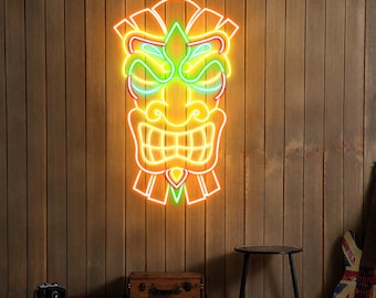 May include: A neon sign in the shape of a Tiki mask, with orange, green, and red lights, is mounted on a wooden wall. The mask has a smiling mouth and detailed facial features. The background includes a stool, luggage, and a guitar.