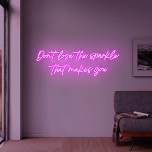 Pode incluir: Uma placa de néon rosa brilhante em uma parede roxa diz: "Don't lose the sparkle that makes you" em uma fonte cursiva. Uma imagem emoldurada e um sofá cinza com uma manta são vistos ao fundo.