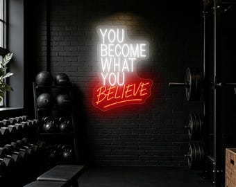 May include: A wall-mounted neon sign featuring the white text "YOU BECOME WHAT YOU" and the red text "BELIEVE" in a cursive script. The sign is mounted on a dark brick wall within a gym setting, illuminated against the dark background.