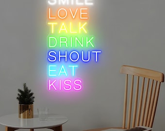 May include: A neon sign with the words "SMILE LOVE TALK DRINK SHOUT EAT KISS" in a vertical arrangement. The sign has a gradient of colors, starting with white at the top and transitioning to pink at the bottom. A small table with a plant and a glass of water is in the foreground.