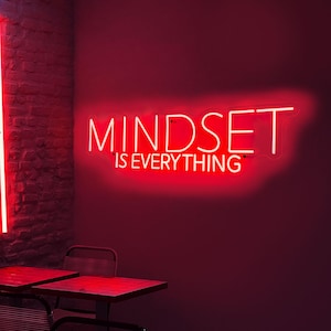 Può includere: Un'insegna al neon rossa su un muro rosso con la scritta "MINDSET IS EVERYTHING". L'insegna è in un font moderno. L'immagine mostra anche un muro di mattoni rossi e un tavolo con sedie.