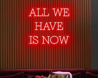 May include: A red neon sign displays the words "ALL WE HAVE IS NOW" against a wooden slatted wall. Below, a silver ice bucket with a bottle and two champagne flutes sit on a burgundy velvet sofa.
