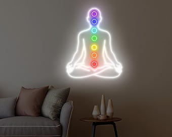 May include: A neon sign depicting a meditating figure with seven colored circles aligned along the spine, representing chakras. The sign is white with a rainbow of colors, including purple, blue, green, yellow, orange, and red. The sign is mounted on a neutral-colored wall.
