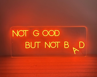 May include: A rectangular neon sign with the words "NOT GOOD BUT NOT BAD" in bright yellow. The sign is lit against an orange background, creating a vibrant and eye-catching display.