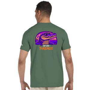 Könnte beinhalten: Ein olivgrünes Kurzarm-T-Shirt mit einer Grafik auf der Rückseite, die ein violettes und orangefarbenes Krokodil mit Sonnenbrille zeigt. Die Grafik enthält einen Sonnenuntergangshintergrund und den orangefarbenen Schriftzug "TOP END THREADS".