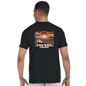 Op de afbeelding: Zwart T-shirt met een woestijnlandschap. Een witte terreinwagen rijdt over een weg in de richting van een zonsondergang. De tekst "TOP END THREADS" staat eronder.