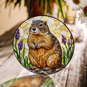 May include: A stained glass window hanging features a brown and tan prairie dog with purple flowers and green leaves. The round window is framed in a dark metal.