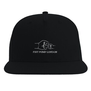 May include: Black baseball cap with a flat peak. The cap has a white embroidered design of two fists bumping, with the text "FIST PUMP LONDON" underneath. A classic cap style, suitable for casual wear.