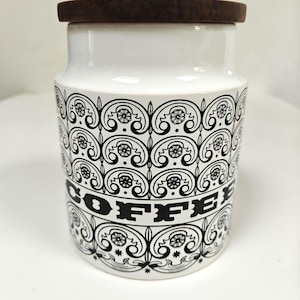 May include: White ceramic coffee canister with a dark wooden lid. The canister features a black and white decorative pattern and the word "COFFEE" in bold letters. A kitchen storage container.