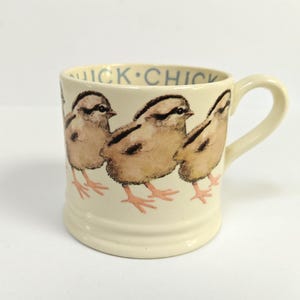 May include: A cream-coloured ceramic mug featuring a repeating watercolour illustration of three fluffy brown chicks. The interior rim of the mug displays the text "QUICK CHICK" in blue lettering. The mug has a sturdy handle and a wide base.