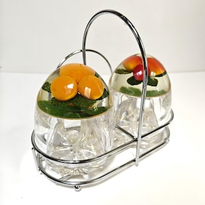 May include: A clear glass salt and pepper shaker set with a chrome metal handle and base. Each shaker has a fruit design inside, with oranges and leaves in one and apples in the other.