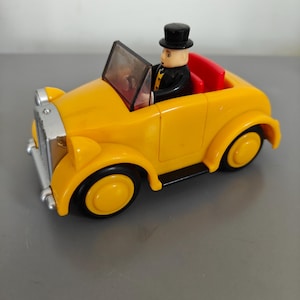 May include: A bright yellow toy convertible car with black wheels and a silver front. A figure in a black suit and top hat sits inside the car. The car's interior is red.