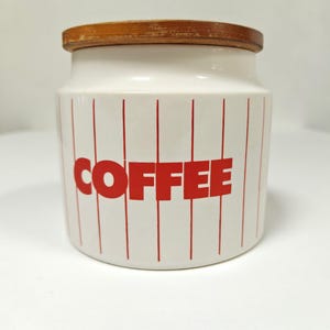 May include: A white ceramic coffee canister with a wooden lid. The canister has vertical red stripes and the word "COFFEE" in large red letters. A kitchen storage container.