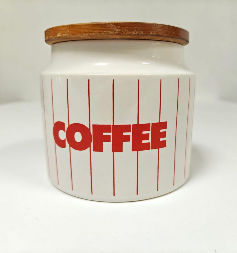 May include: A white ceramic coffee canister with a wooden lid. The canister has vertical red stripes and the word "COFFEE" in large red letters. A kitchen storage container.