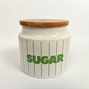 May include: A white ceramic storage canister with a cylindrical shape, featuring vertical green pinstripes and the word SUGAR printed in bold green capital letters on the front. The container is topped with a round, natural light brown wooden lid.