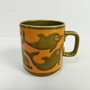 Może przedstawiać: Ceramiczny kubek z żywym pomarańczowym tłem ozdobionym wieloma ciemnooliwkowymi sylwetkami delfinów. Kubek posiada pasujący oliwkowy brzeg, solidny uchwyt oraz cylindryczny korpus z gładkim, szkliwionym wykończeniem na całej powierzchni.