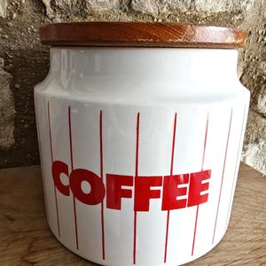 May include: White ceramic coffee canister with red vertical stripes and the word "COFFEE" in red block letters. The canister has a wooden lid and is sitting on a wooden surface.