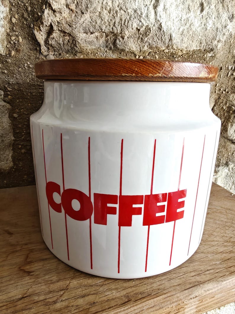 May include: White ceramic coffee canister with red vertical stripes and the word "COFFEE" in red block letters. The canister has a wooden lid and is sitting on a wooden surface.