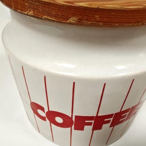 May include: White ceramic coffee canister with a wooden lid. The canister has red vertical lines and the word "COFFEE" in red block letters. The lid is made of light brown wood.