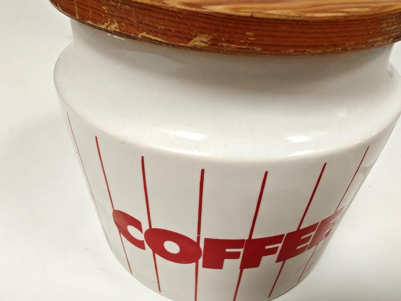May include: White ceramic coffee canister with a wooden lid. The canister has red vertical lines and the word "COFFEE" in red block letters. The lid is made of light brown wood.