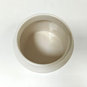 May include: A cream-colored ceramic bowl with a rounded shape. The bowl has a smooth, glossy finish and a wide opening. The interior of the bowl is a lighter shade of cream. The bowl is likely used for serving or storage.
