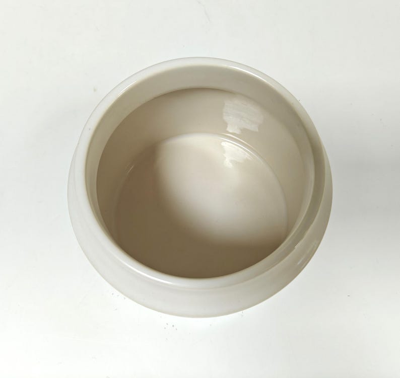 May include: A cream-colored ceramic bowl with a rounded shape. The bowl has a smooth, glossy finish and a wide opening. The interior of the bowl is a lighter shade of cream. The bowl is likely used for serving or storage.