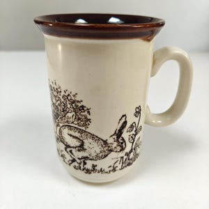Könnte beinhalten: Cremefarbene Keramiktasse mit braunem Rand und Henkel. Die Tasse zeigt eine detaillierte schwarze Illustration eines Hasen in einer natürlichen Umgebung. Ein klassisches Design für Tee oder Kaffee.