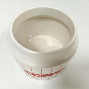 May include: A white ceramic coffee canister with red lettering and vertical lines. The canister has a wide opening and a rounded shape. The interior is a glossy white. The word "COFFEE" is visible on the side.