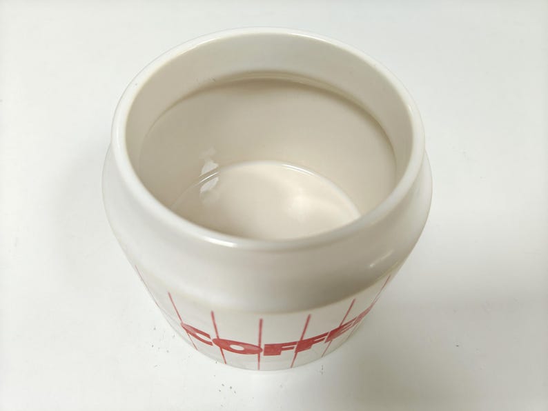 May include: A white ceramic coffee canister with red lettering and vertical lines. The canister has a wide opening and a rounded shape. The interior is a glossy white. The word "COFFEE" is visible on the side.