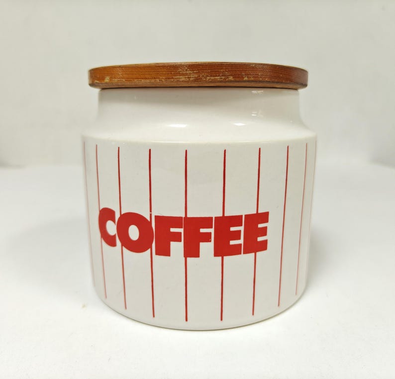May include: A white ceramic coffee canister with a wooden lid. The canister has red vertical stripes and the word "COFFEE" in large red letters. A kitchen storage container.