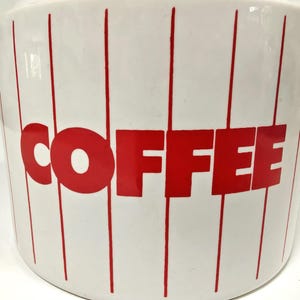 May include: White ceramic coffee canister with red vertical stripes and the word "COFFEE" in large red letters. The canister has a rounded shape and a glossy finish, perfect for kitchen storage.