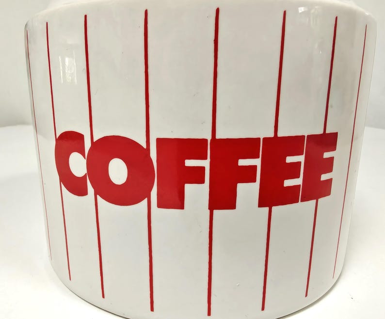 May include: White ceramic coffee canister with red vertical stripes and the word "COFFEE" in large red letters. The canister has a rounded shape and a glossy finish, perfect for kitchen storage.